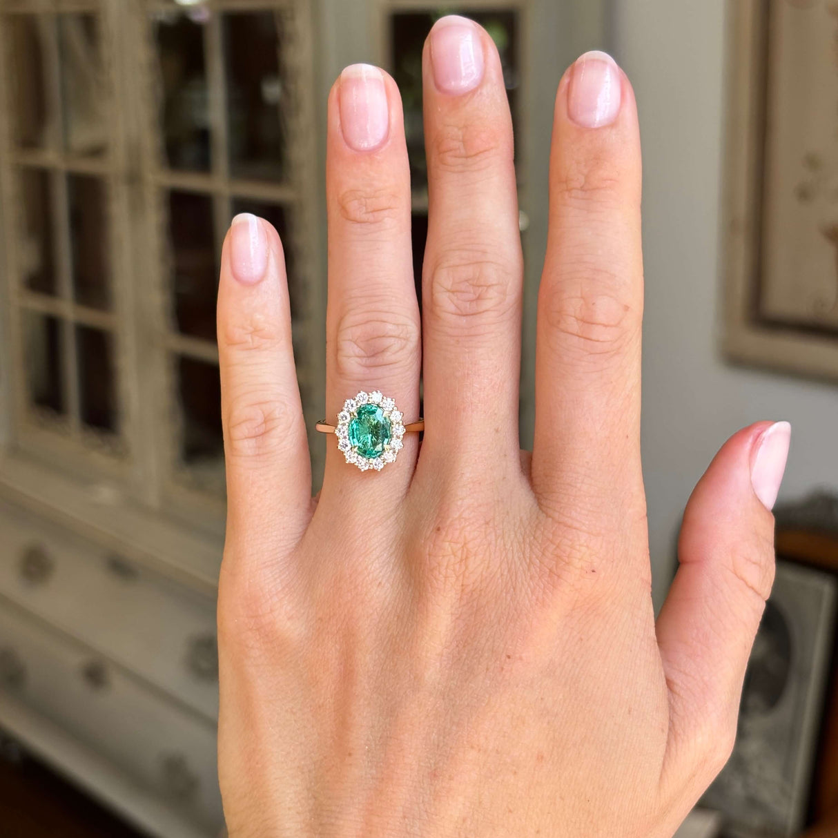 Antique-inspired yellow gold emerald and diamond cluster ring worn on hand, featuring an oval-cut emerald centre stone with a brilliant diamond halo in natural light.