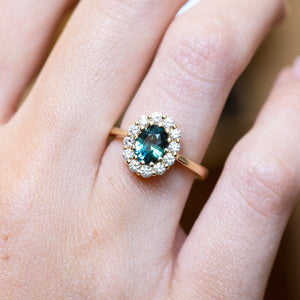 Green sapphire and diamond cluster engagement ring with oval-cut sapphire and diamond halo worn on hand.