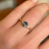 Oval green sapphire and diamond trilogy engagement ring in yellow gold worn on hand.