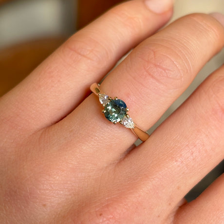 Oval green sapphire and diamond trilogy engagement ring in yellow gold worn on hand.