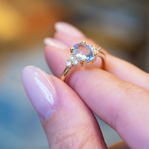 Pale blue sapphire and diamond cluster tre-foil engagement ring held between fingers showing diamond cluster detail.
