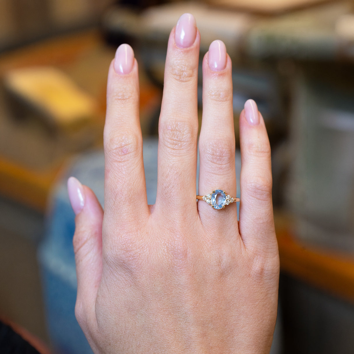 Pale blue sapphire and diamond cluster tre-foil engagement ring worn on hand with oval sapphire centre stone.
