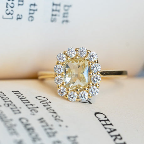 Pale yellow sapphire and diamond cluster engagement ring with surrounding diamond halo on book pages.