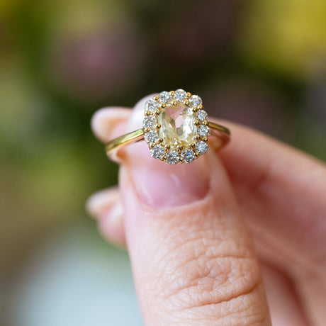 Pale yellow sapphire and diamond cluster engagement ring held between fingers with diamond surround.