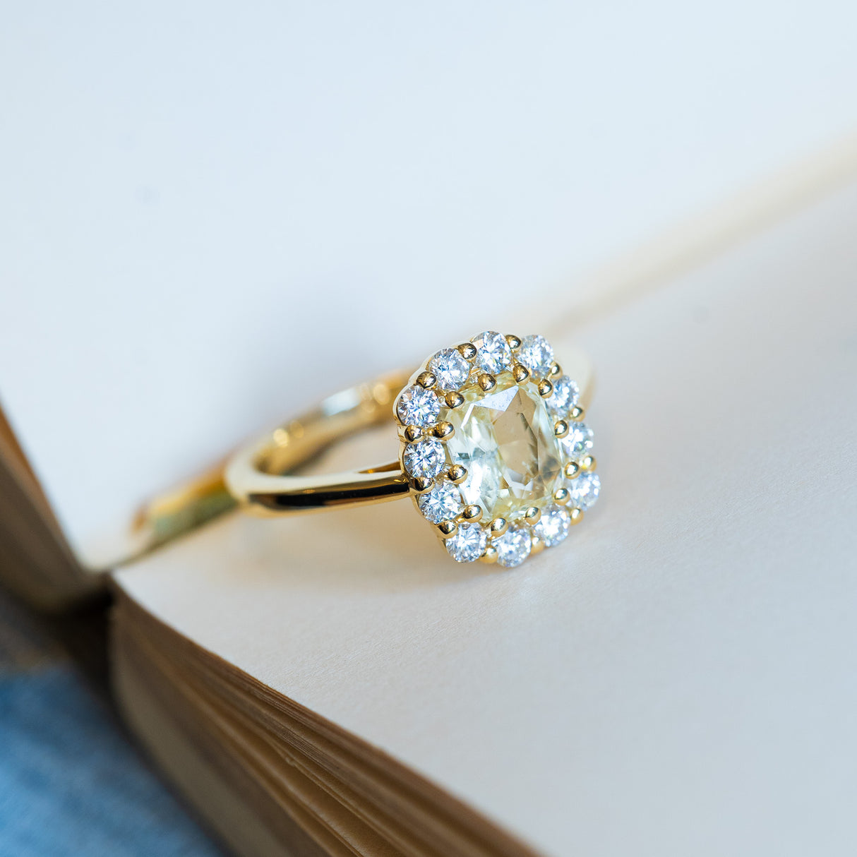 Pale yellow sapphire and diamond cluster engagement ring resting on open book pages.