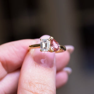 Peach coloured sapphire and diamond Toi et Moi engagement ring held between fingers with emerald cut diamond and pear sapphire.