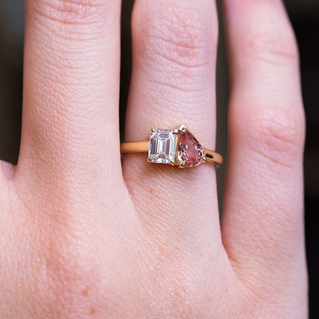 Peach coloured sapphire and diamond Toi et Moi engagement ring with emerald cut diamond and pear sapphire worn on finger.