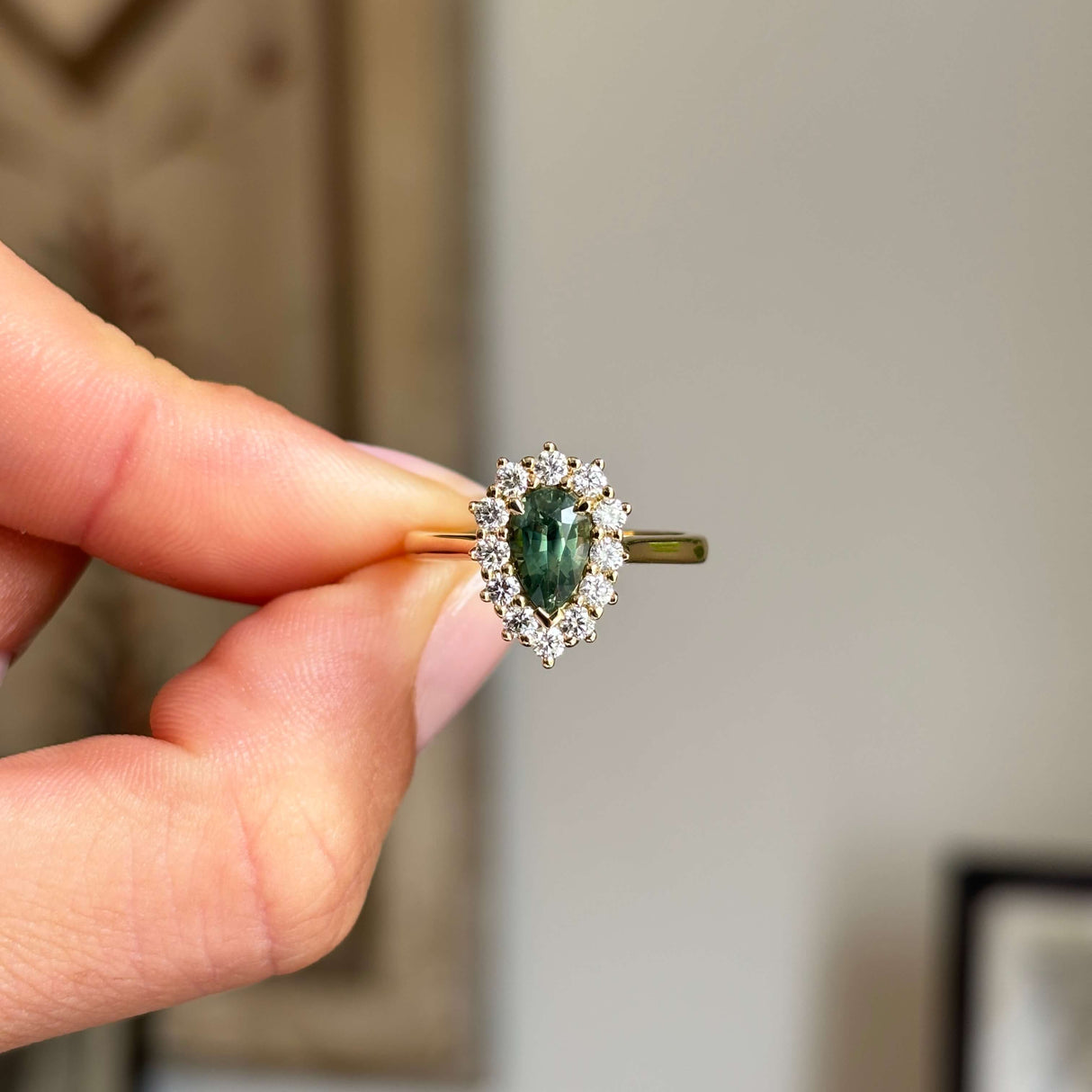 Antique-inspired pear-cut green sapphire surrounded by brilliant diamonds in a yellow gold cluster setting, held between fingers against a softly blurred vintage interior background.