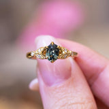 Pine green sapphire and diamond engagement ring held between fingers with gold band.
