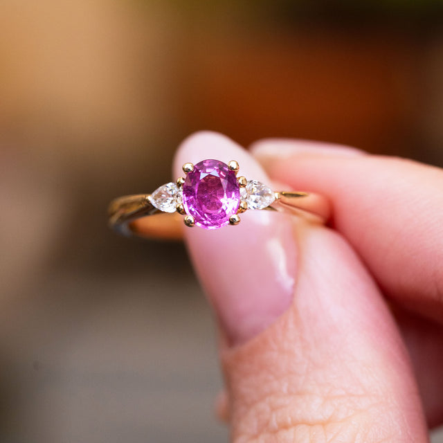 Pink oval sapphire and diamond three-stone engagement ring held between fingers with gold band.