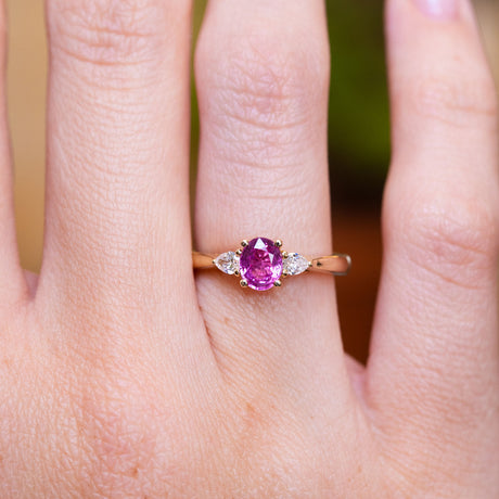 Pink oval sapphire and diamond three-stone engagement ring worn on finger with oval centre stone.