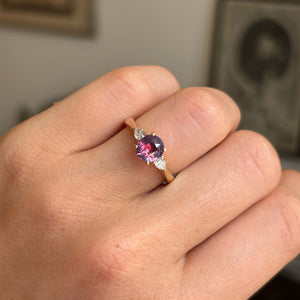 Pink-red sapphire and diamond three-stone engagement ring featuring a vibrant pink-red sapphire flanked by pear-cut diamonds, worn on a hand.