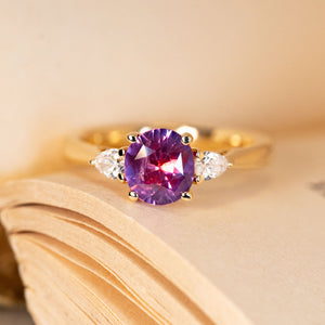 Pink-red sapphire and diamond three-stone engagement ring featuring a vibrant oval sapphire flanked by pear-cut diamonds in yellow gold.