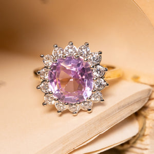 Engagement ring showcasing a soft pink Ceylon sapphire encircled by brilliant-cut diamonds, creating an elegant vintage-inspired cluster design.