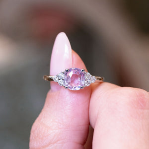 Pink sapphire and diamond cluster tre-foil engagement ring held between fingers with oval pink sapphire centre stone.