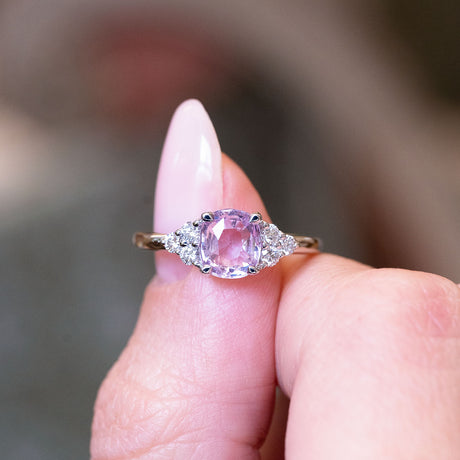 Pink sapphire and diamond cluster tre-foil engagement ring held between fingers with oval pink sapphire centre stone.
