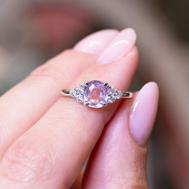 Pink sapphire and diamond cluster tre-foil engagement ring with diamond side clusters held between fingers.