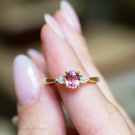 Pink sapphire and diamond three-stone engagement ring held between fingers.