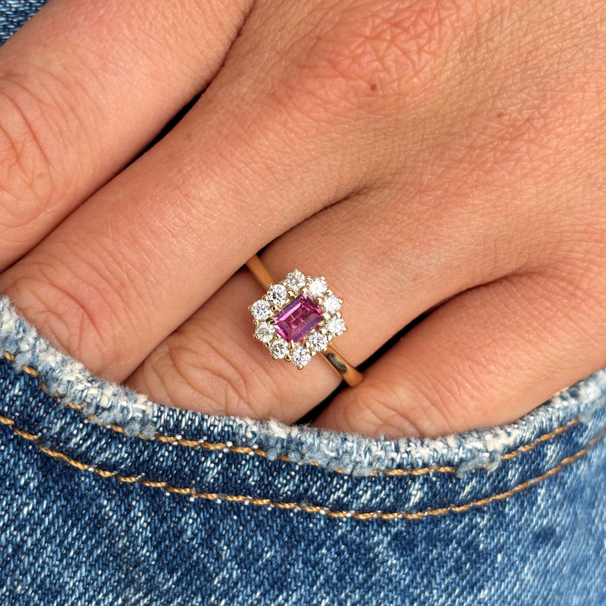 Antique-inspired yellow gold diamond cluster ring with a step-cut pinkish-red ruby centre stone, styled on hand against blue denim to showcase its vivid colour and elegant design.