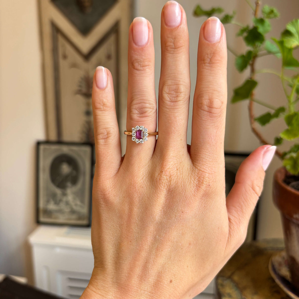 Antique-inspired yellow gold ruby and diamond cluster ring worn on hand, featuring a vibrant step-cut pinkish-red ruby framed by sparkling diamonds in natural light.