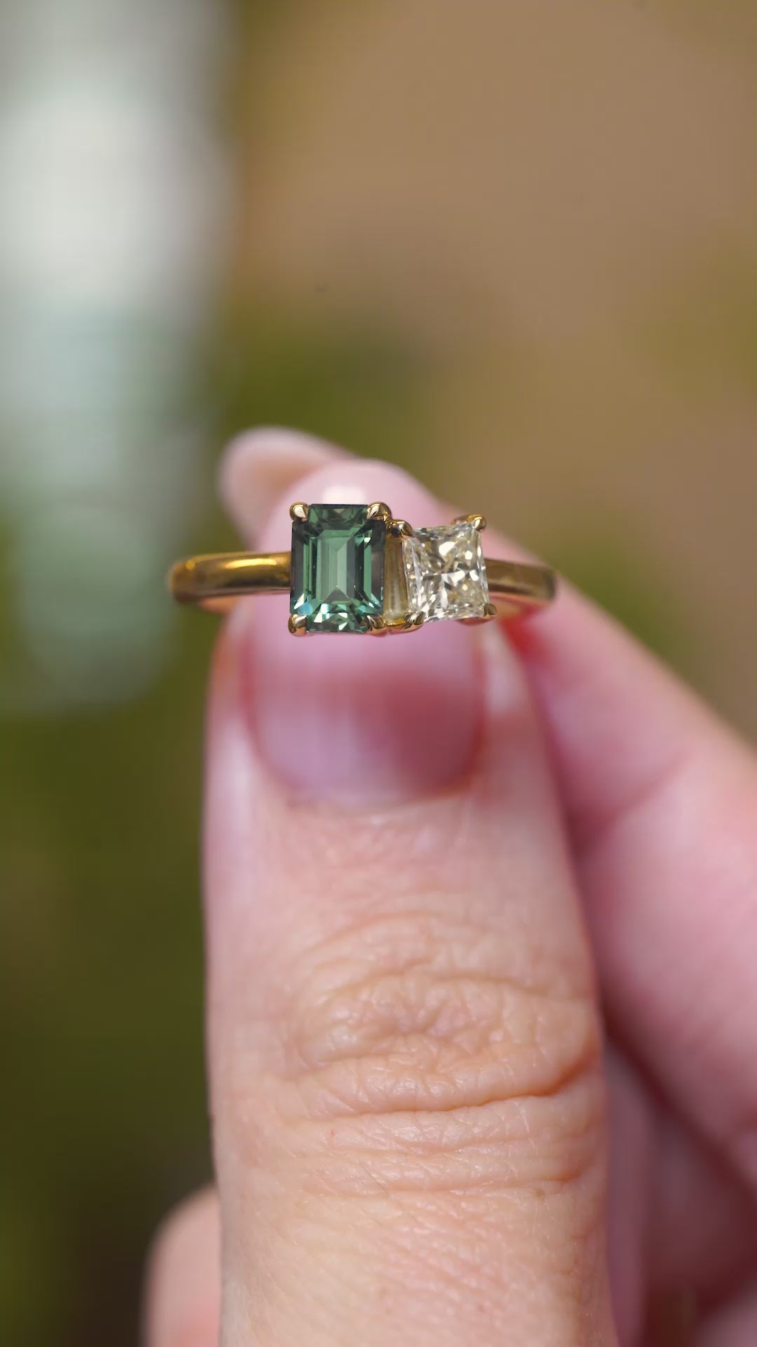 A video of a toi et moi green sapphire and diamond engagement ring held between fingers.