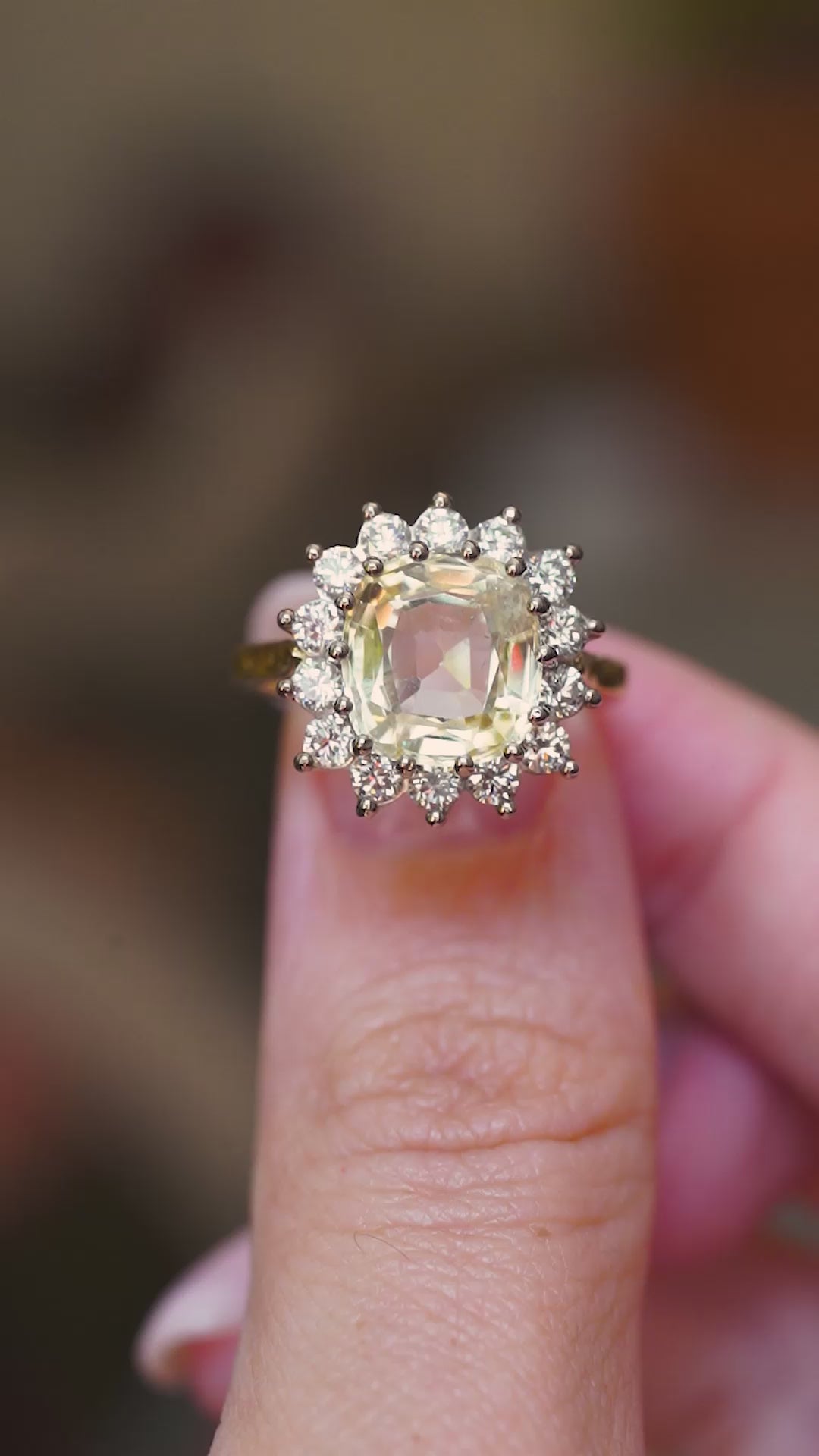 A vintage-inspired, yellow Ceylon sapphire and diamond cluster engagement ring shown between fingers in video.
