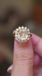 A vintage-inspired, yellow Ceylon sapphire and diamond cluster engagement ring shown between fingers in video.