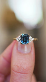 Sapphire and diamond ring showing central teal stone and surrounding sparkle detail between fingers.