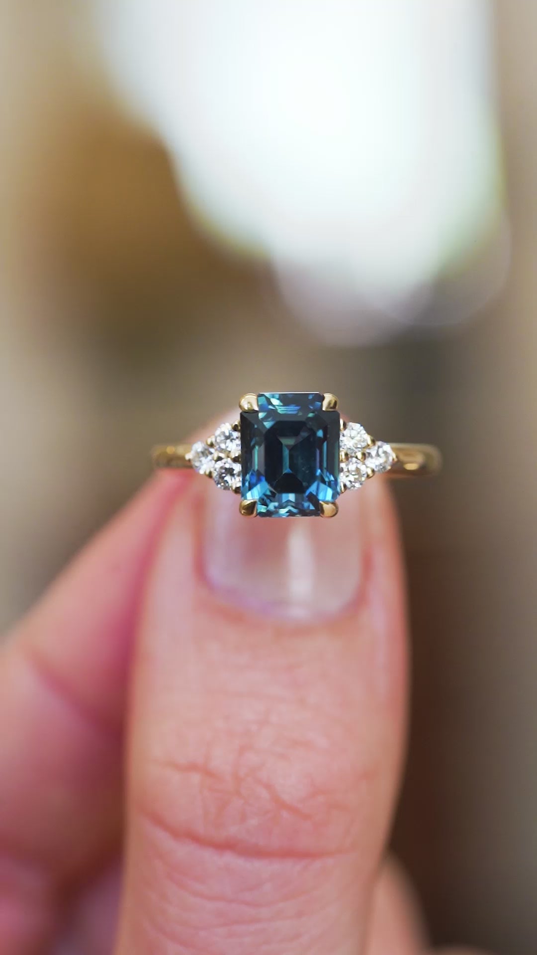 Sapphire and diamond ring showing central teal stone and surrounding sparkle detail between fingers.