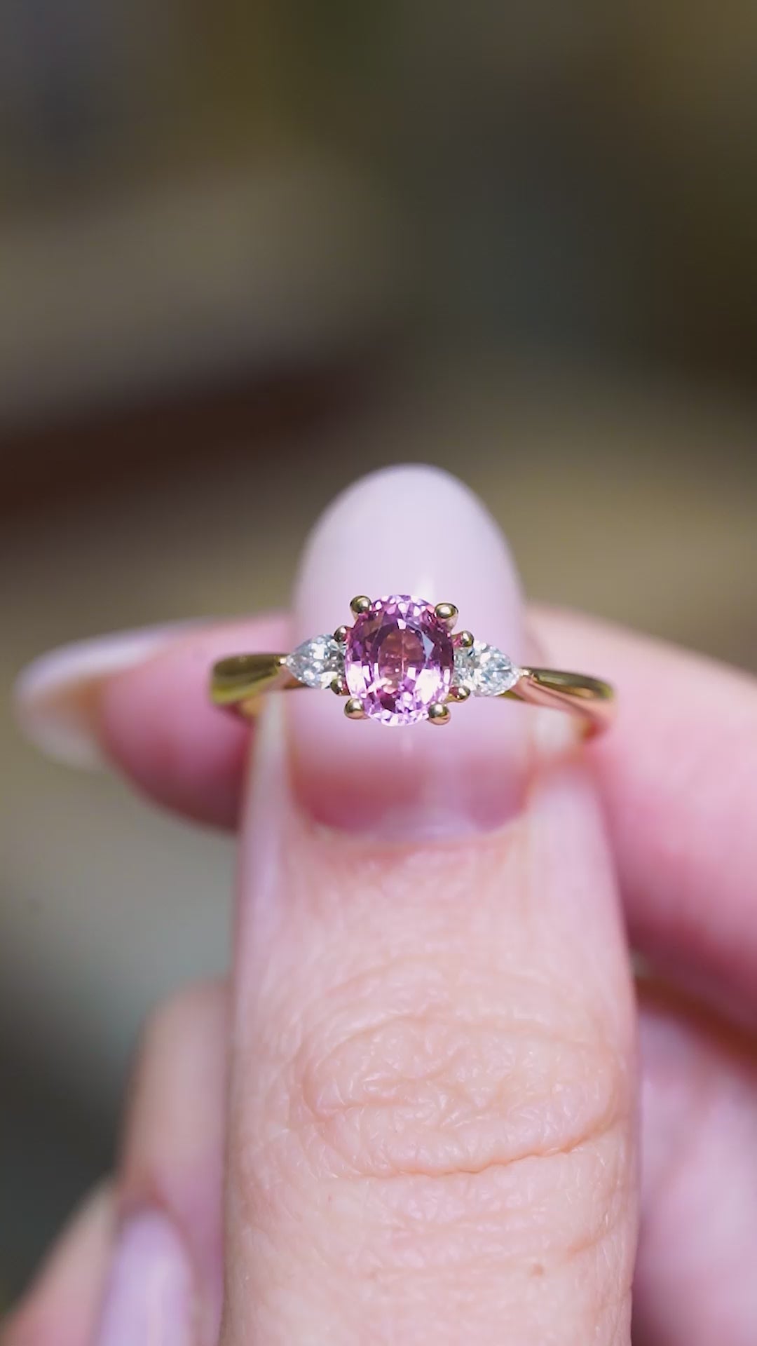 Pink sapphire and diamond three-stone engagement ring held between fingers showing ring details.