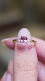 Pink sapphire and diamond three-stone engagement ring held between fingers showing ring details.