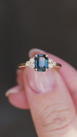 A close-up of a yellow gold ring with a teal sapphire and diamond cluster.