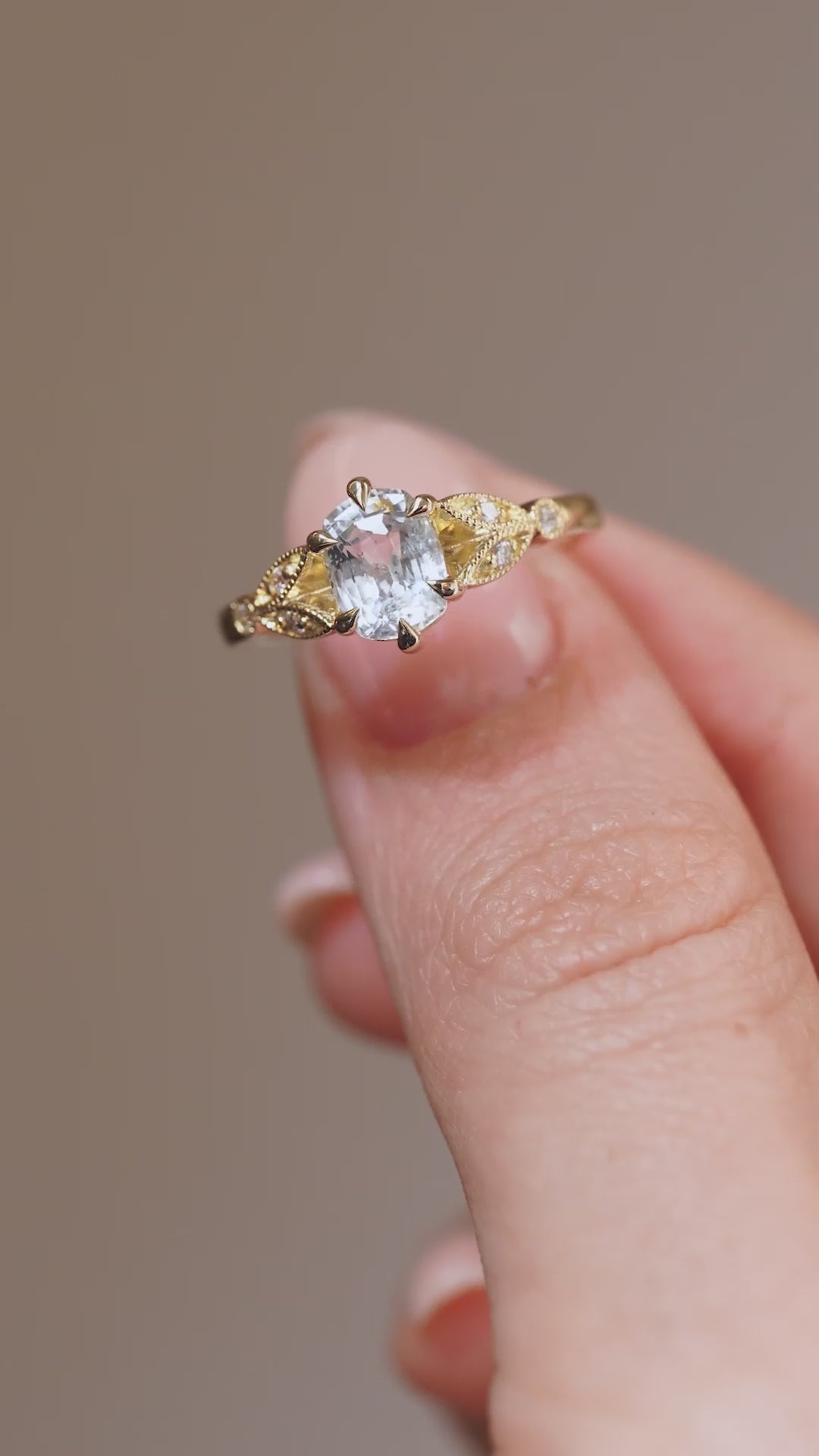 A close-up video of the engagement ring held between fingers, showcasing the sparkling white sapphire and detailed gold craftsmanship.