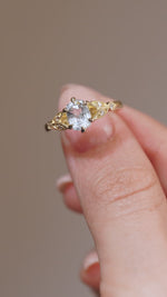 A close-up video of the engagement ring held between fingers, showcasing the sparkling white sapphire and detailed gold craftsmanship.
