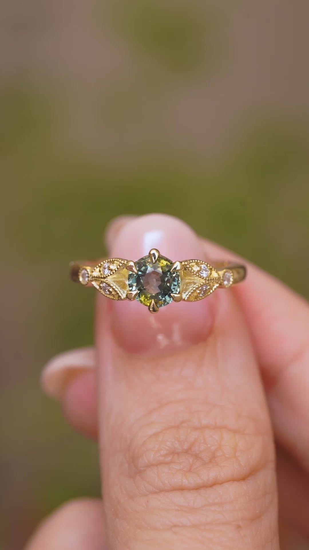 A close up video of a Vintage-inspired, Green sapphire and diamond engagement ring in yellow gold held between two fingers and rotated slowly to display all the profiles of the ring.