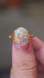 A video of an antique, Edwardian era oval opal single stone ring with decorative split shoulders mounted in yellow gold and held between the fingers as it is turned to show the different angles and details of the ring.