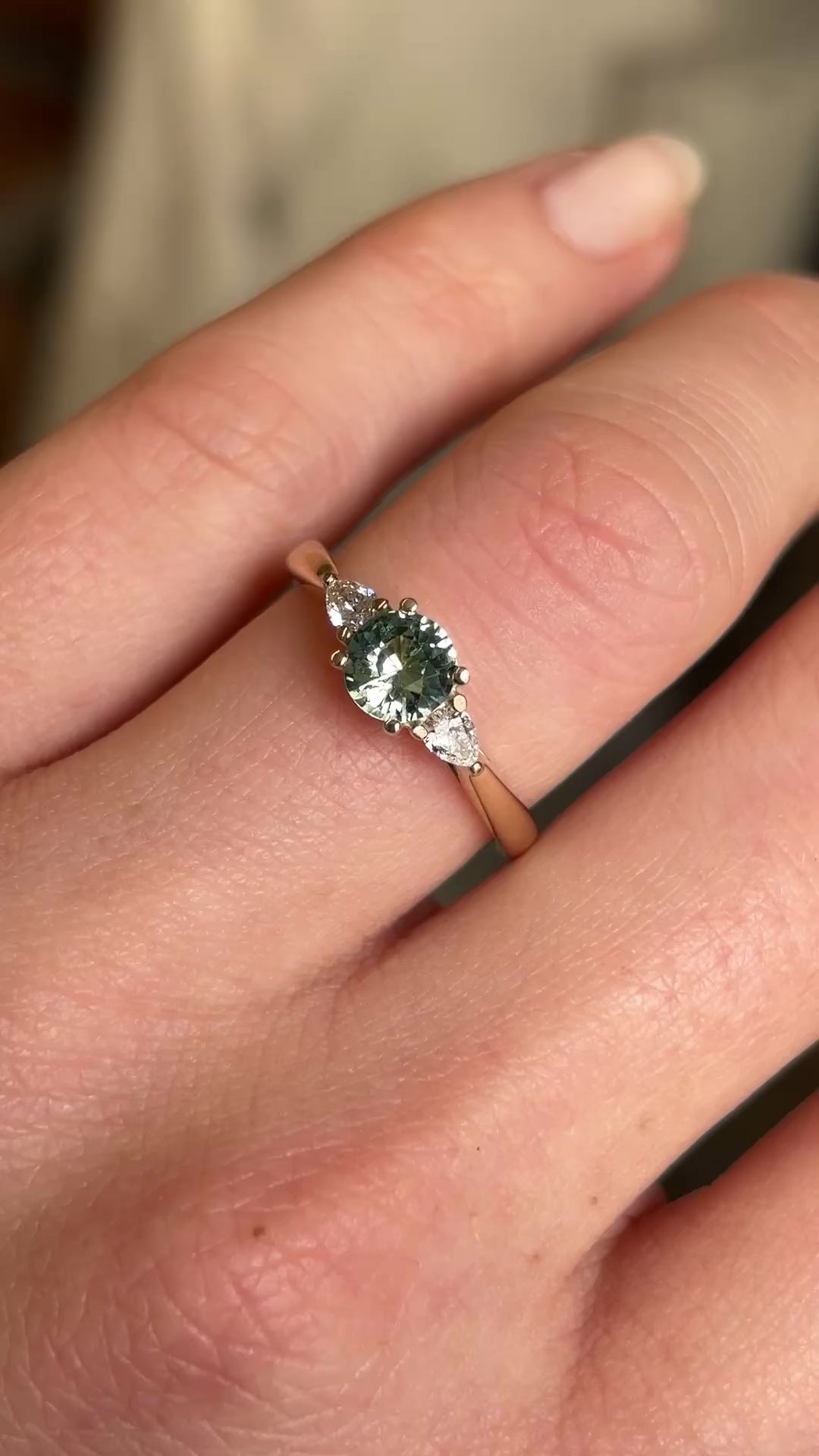 Round teal sapphire and pear-cut diamond three-stone engagement ring worn on the hand, highlighting the sapphire’s colour and pear-cut diamonds in natural light near a window.