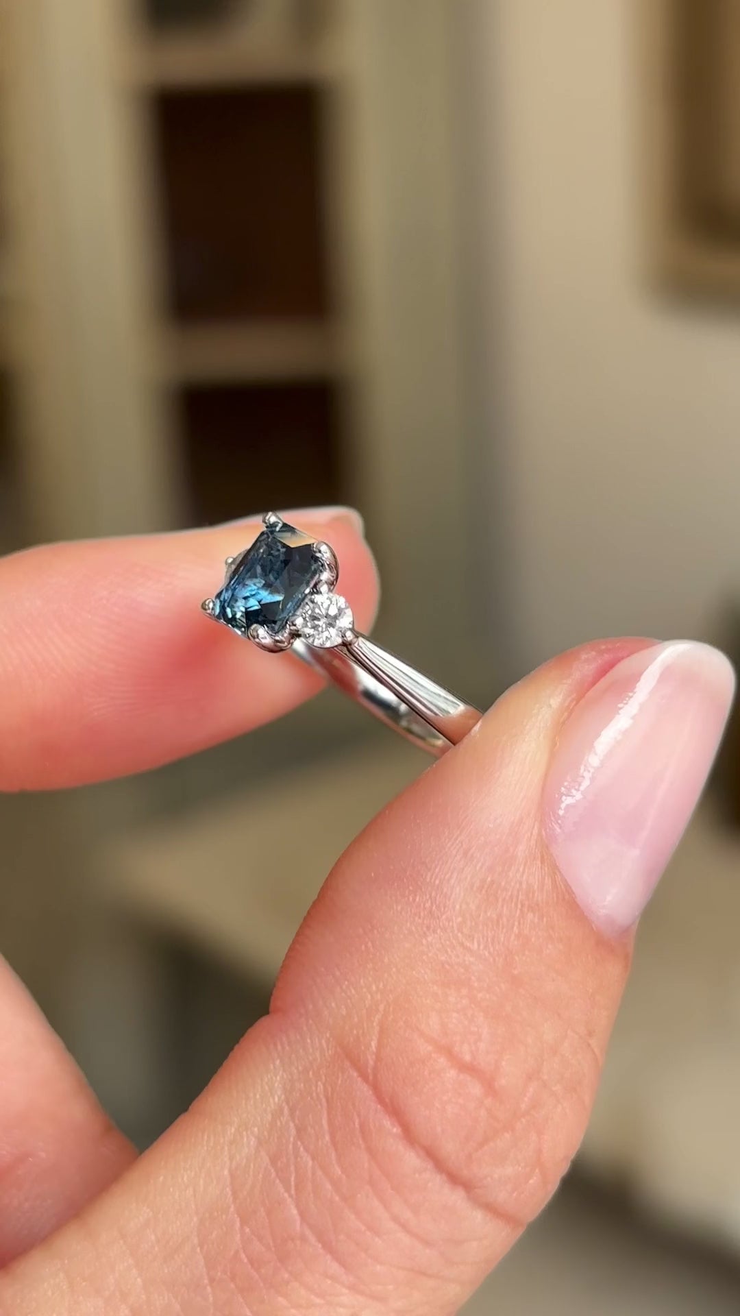 Video of a Vintage-inspired, Teal sapphire and diamond three-stone engagement ring in platinum held by two fingers and rotated to show the profile