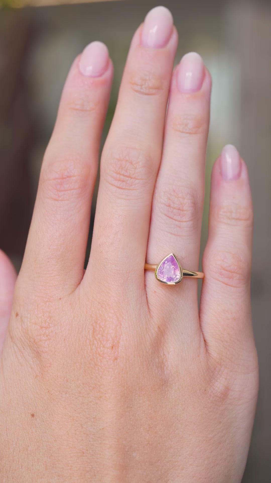 A video of a hand wearing an antique-inspired, light Pink sapphire solitaire engagement ring, the hand is stretched out and slowly turned against a blurred background.