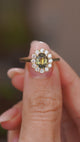 A close up video of an Antique-inspired, Green oval sapphire and diamond cluster engagement ring held between two fingers and rotated slowly to display the yellow gold wedding fit setting and band, against a softly blurred background. 