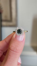 An Antique-inspired, teal sapphire and diamond cluster engagement ring being shown in video held between two fingers.