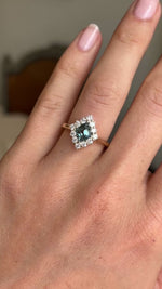 A video of an Antique-inspired, Teal kite-cut sapphire and diamond cluster engagement ring in yellow gold worn on a wedding finger against a blurred background