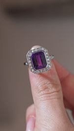 A close-up video of an antique Art Deco amethyst and diamond cluster ring held between fingers, showcasing the gemstone's deep purple colour, step-cut facets and diamond-set geometric surround.
