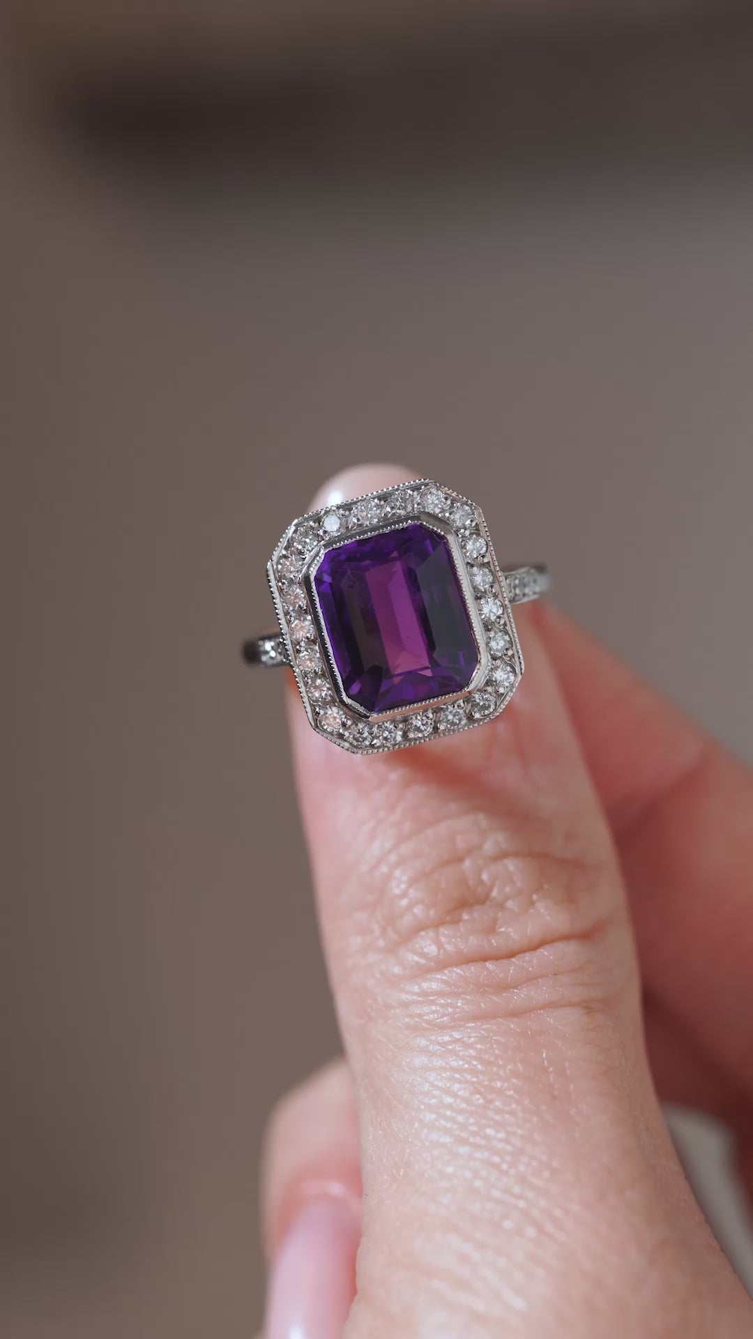 A close-up video of an antique Art Deco amethyst and diamond cluster ring held between fingers, showcasing the gemstone's deep purple colour, step-cut facets and diamond-set geometric surround.