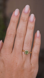 Close up video of an Antique-inspired, Green sapphire solitaire engagement ring worn on a wedding finger and rotated slowly to show the profile of the yellow gold setting and band once worn.  Against a soft interior background. 