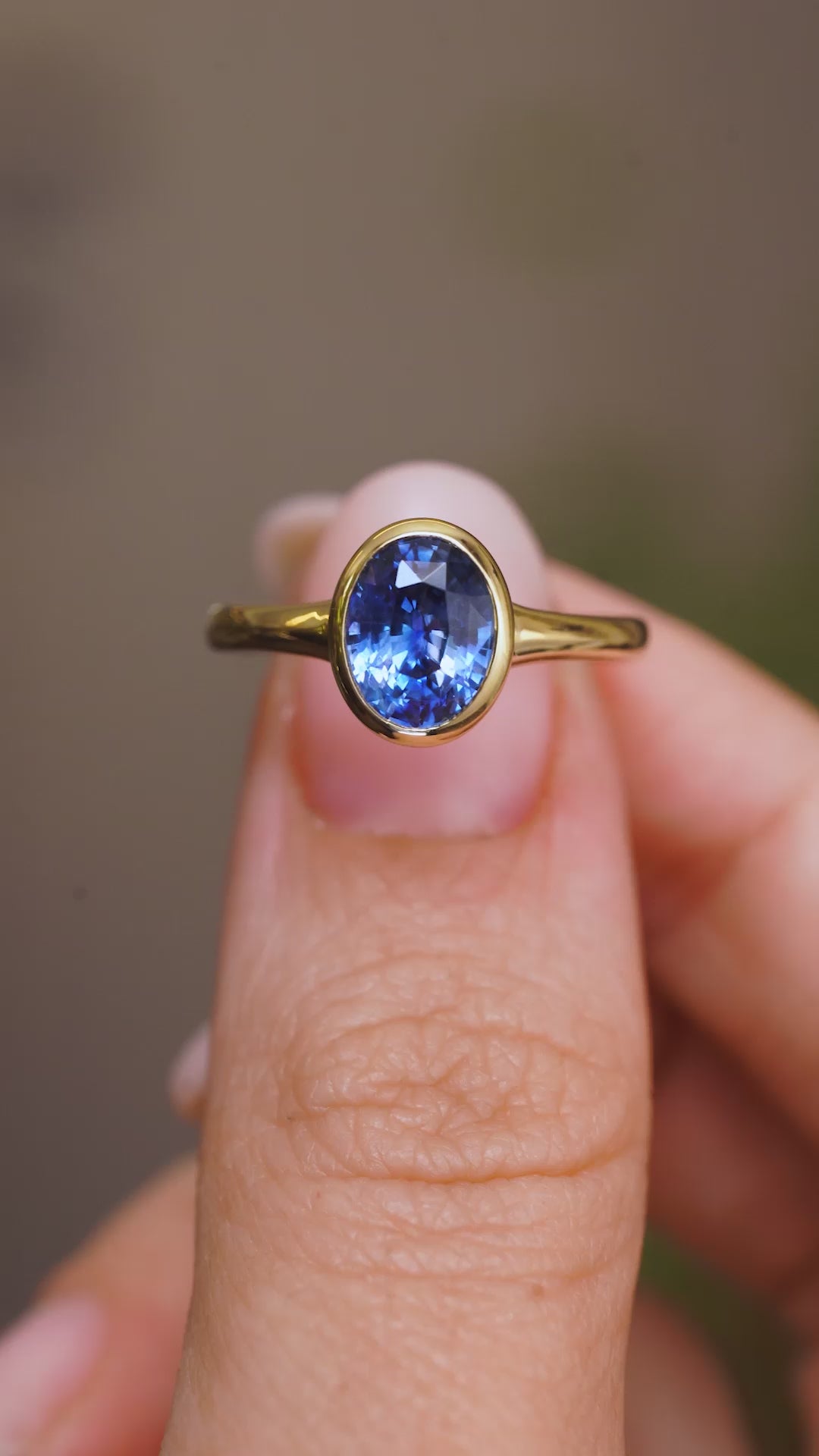 A close up video of a Vintage-inspired blue sapphire solitaire engagement ring, held between two fingers and rotated slowly to showcase the yellow gold rub-over setting of the Sri Lankan sapphire and the yellow gold band.  