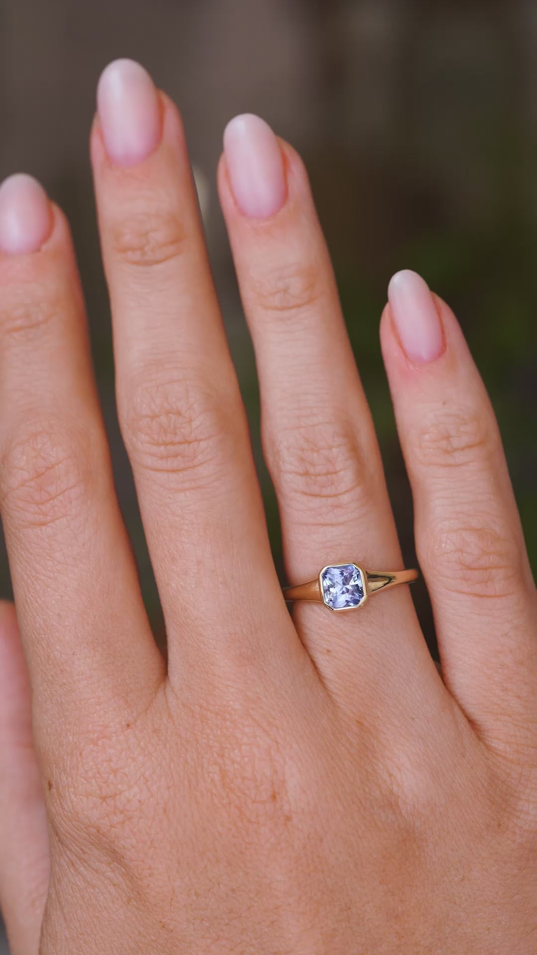 A video of a solitaire engagement ring worn on a wedding finger and rotated slowly to show the Antique-inspired rub-over setting design, the blue square-cut sapphire and the yellow gold band.  