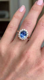 Antique-inspired intense cornflower blue sapphire and diamond cluster engagement ring featuring an oval cornflower blue sapphire surrounded by brilliant-cut diamonds, displayed on a hand.