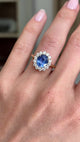 Antique-inspired intense cornflower blue sapphire and diamond cluster engagement ring featuring an oval cornflower blue sapphire surrounded by brilliant-cut diamonds, displayed on a hand.