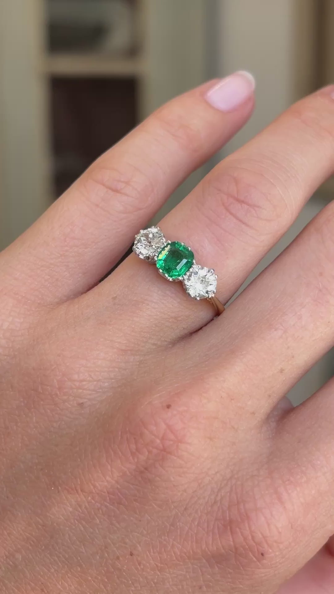 Antique Edwardian emerald and diamond three-stone engagement ring featuring an emerald centre stone flanked by two diamonds, displayed on a hand.